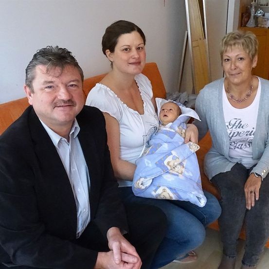 Drei Erwachsene und ein Baby sitzen auf einer orangefarbenen Couch und lächeln für die Kamera. Das Baby ist in eine blaue Decke gewickelt.