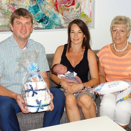 Eine dreiköpfige Familie, bestehend aus einem lächelnden Mann, einer Frau, die ein schlafendes Baby hält, und einer älteren Frau, die alle auf einer Couch sitzen. Sie halten ein eingewickeltes Geschenk und ein Kissen.