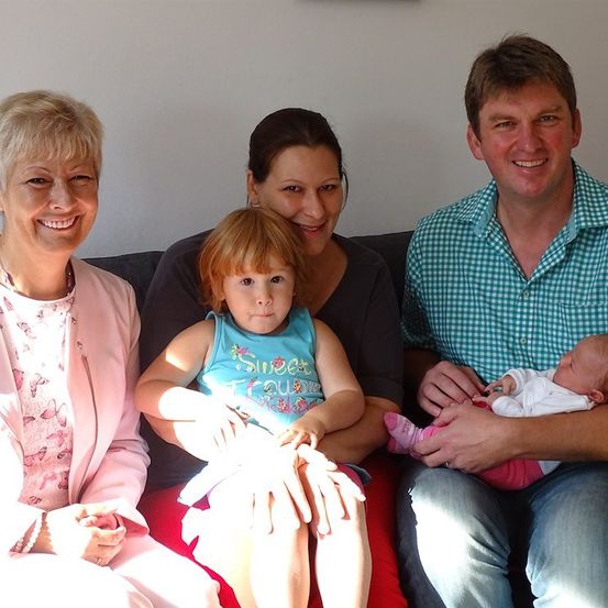 Eine vierköpfige Familie, bestehend aus einer lächelnden Frau, einer Frau, die ein Kleinkind hält, einem Mann, der ein Baby hält, und einer Frau, die auf einer Couch sitzt.