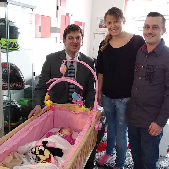 Drei Personen stehen neben einem Baby in einem rosa Kinderbett mit Spielzeug darüber. Der Mann trägt einen Anzug und eine Krawatte. Die Frau trägt ein schwarzes Shirt und pinke Hausschuhe. Der Mann rechts trägt ein graues Shirt. Dahinter steht ein Glasschrank mit Helmen.