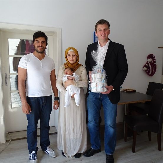 Drei Personen stehen in einem Raum, einer hält ein Baby und der andere ein eingewickeltes Geschenk. Der Mann auf der linken Seite trägt ein weißes T-Shirt und Jeans, die Frau in der Mitte trägt einen Hijab und hält ein Baby, und der Mann auf der rechten Seite trägt einen Anzug und hält ein eingewickeltes Geschenk. Hinter ihnen steht eine Tür, ein Tisch und ein Stuhl.