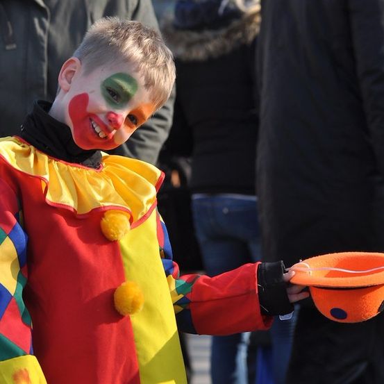 Ein junger Junge, der als Clown verkleidet ist, hält einen kleinen orangefarbenen Hut und lächelt fröhlich in die Kamera. Er trägt ein farbenfrohes Kostüm mit gelbem Kragen.