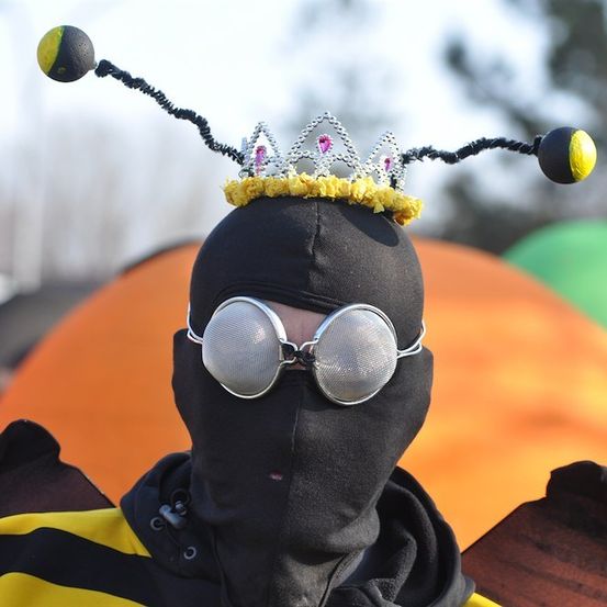 Eine Person in einem Bienenkostüm mit Krone, schwarzem Maske und silbernen Schutzbrillen. Der Hintergrund ist verschwommen mit orangen und grünen Farben.