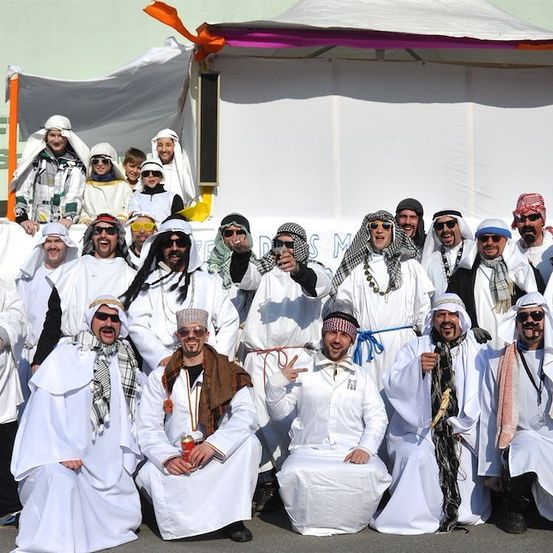 Eine Gruppe von Menschen in weißen Roben und Kopftüchern, die vor einem Zelt für ein Foto posieren. Einige halten Getränke und tragen Sonnenbrillen.