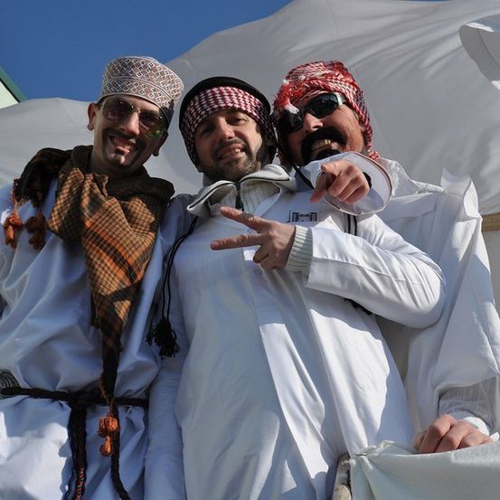 Drei Männer in weißen Roben und traditioneller Kopfbedeckung, einer mit einem Schal, posieren für ein Foto vor einem weißen Zelt.