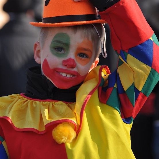 Ein junger Junge, der als Clown verkleidet ist, mit bunter Gesichtsbemalung und Hut, lächelt in die Kamera.
