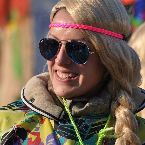 Eine Frau mit blonden Haaren, die eine rosa Kopfband, Sonnenbrille und eine bunte Jacke mit einem grünen Seil um den Hals trägt. Sie lächelt und scheint sich bei einer Veranstaltung im Freien zu befinden.