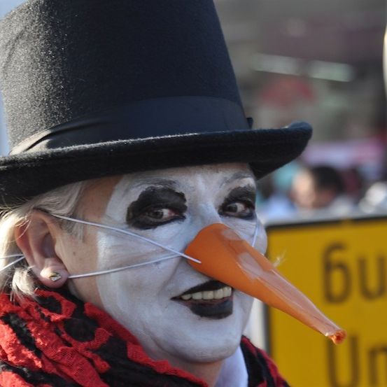 Eine Person in einem Clownkostüm mit einer großen orangefarbenen Nase und einem schwarzen Zylinder steht vor einem verschwommenen Hintergrund.