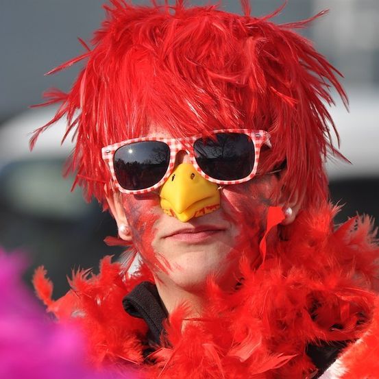 Eine Person mit roten Federn um den Hals und Gesichtsbemalung auf dem Gesicht, trägt eine rote Perücke und eine Sonnenbrille.