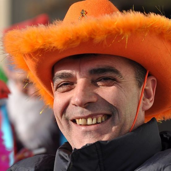Ein Mann mit einem orangefarbenen Hut mit einer dekorativen Brosche lächelt in die Kamera.