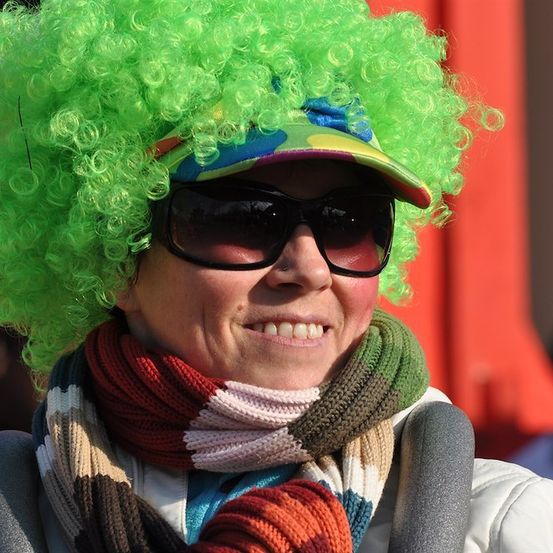 Eine Frau mit grünen lockigen Haaren, die eine bunte Mütze, Sonnenbrille und einen mehrfarbigen Schal trägt, lächelt in die Kamera.