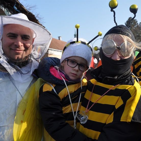 Drei Personen in Bienenkostümen, einer mit einem Imkerhut, einer mit einer Maske und einem Kind dazwischen. Sie lächeln und posieren für ein Foto.