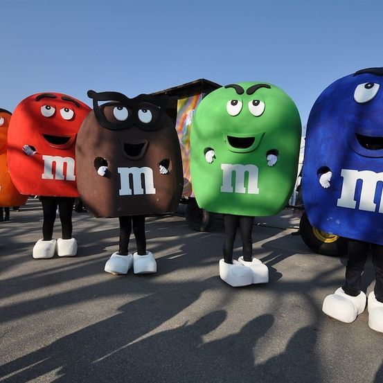 Fünf Maskottchen in M&M's-Kostümen stehen in einer Reihe, jedes repräsentiert eine andere Farbe der Süßigkeit. Sie tragen weiße Stiefel und stehen auf einer gepflasterten Oberfläche.