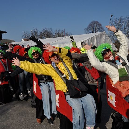 Eine Gruppe von Menschen in bunten Kostümen und Perücken sitzt auf roten und gelben Sitzen, wahrscheinlich für eine Parade. Sie heben die Hände und scheinen Spaß zu haben.