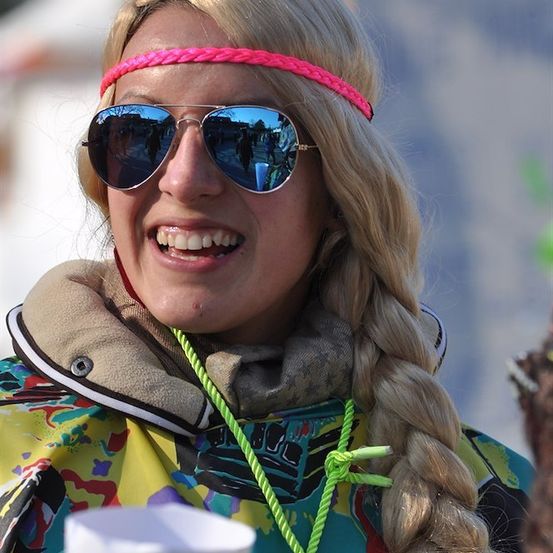 Eine Frau mit blonden Haaren trägt eine rosa Kopfband, Sonnenbrille und eine bunte Jacke und lächelt.