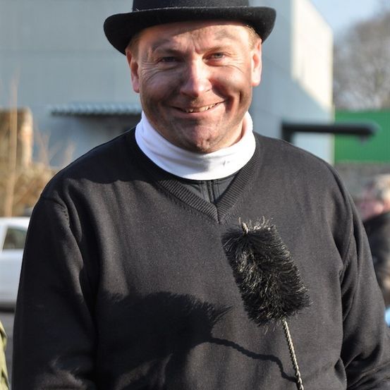 Ein Mann in einem schwarzen Hut und einem schwarzen Pullover lächelt für die Kamera, mit einem schwarzen Federbesen vor ihm.