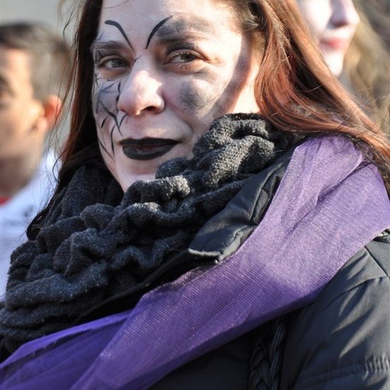 Eine Frau mit dunklem Make-up um die Augen und auf dem Gesicht, trägt einen lila Schal und eine schwarze Jacke.