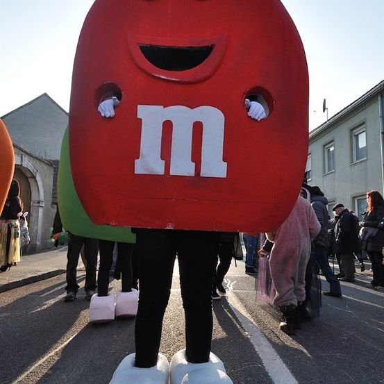 Eine Person in einer M&M-Kostümverkleidung mit einem roten Kostüm und weißen Schuhen steht auf einer Straße mit anderen Leuten im Hintergrund.