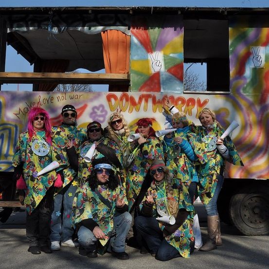 Eine Gruppe von Menschen in farbenfrohen, psychedelischen Kostümen posiert vor einem bunt bemalten Lastwagen. Der Lastwagen hat Graffiti mit Friedenszeichen und den Worten 'Make Love Not War'.