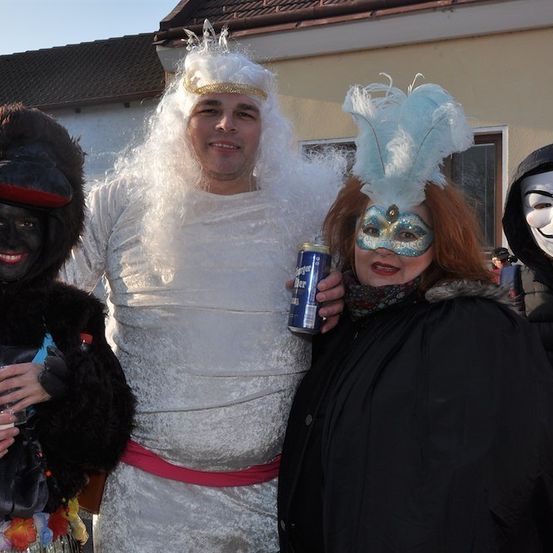 Vier Menschen in Kostümen posieren für ein Foto. Einer hält eine Bierdose. Die Kostüme umfassen einen weißen Umhang, eine schwarze Maske, einen schwarzen Pelzmantel und eine Federmaske.