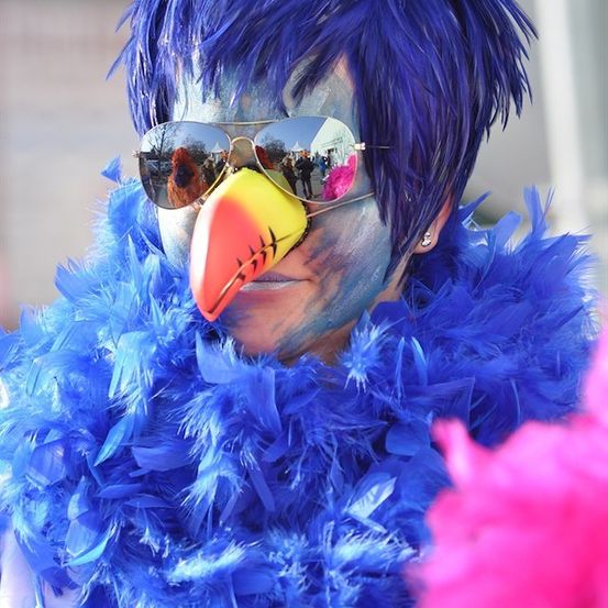 Eine Person mit blauen Haaren, Gesichtsbemalung und einer blauen Federboa, die eine schnabelförmige Maske und Sonnenbrille trägt.