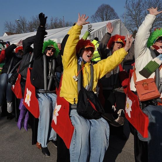 Eine Gruppe von Menschen in bunten Kostümen und Perücken fährt auf einer Achterbahn und jubelt. Sie halten rote Flaggen mit weißen Blumen. Hinter ihnen steht ein weißes Zelt und Bäume.