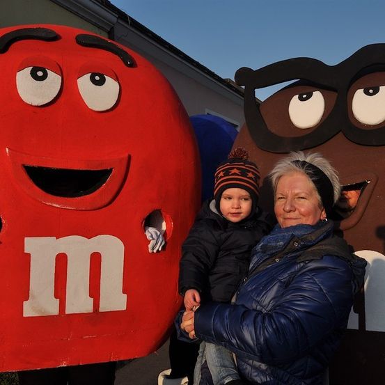 Eine ältere Frau und ein Kind stehen neben großen Kostümen von M&M-Charakteren, einem roten und einem braunen, vor einem Gebäude.