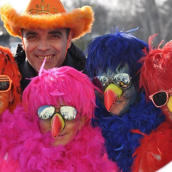 Fünf Personen in bunten Kostümen und Gesichtsbemalung, lächelnd und posierend für ein Foto. Der Mann in der Mitte trägt ein rosa Federkostüm.