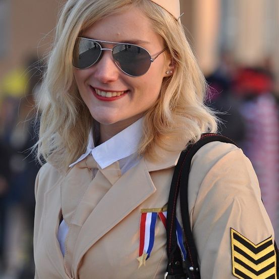 Eine Frau in einer beigen Uniform mit Hut und Sonnenbrille, lächelt und steht im Freien. Sie hat eine Medaille und einen Schulterabzeichen an ihrer Uniform.