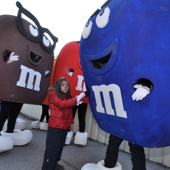 Ein junges Mädchen in einer roten Jacke steht neben großen Maskottchen der M&M's-Figuren in Braun, Rot und Blau. Die Maskottchen haben Löcher für Augen und Münder.