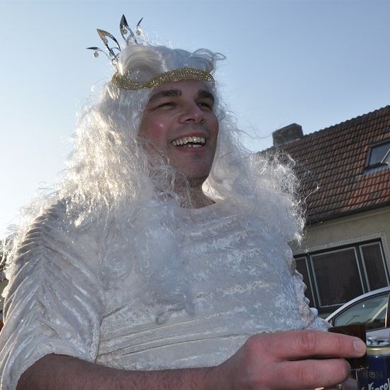Ein Mann in einem weißen Kostüm mit goldener Krone, lächelt und hält eine Bierdose. Dahinter ein Haus mit rotem Dach und ein geparktes Auto.