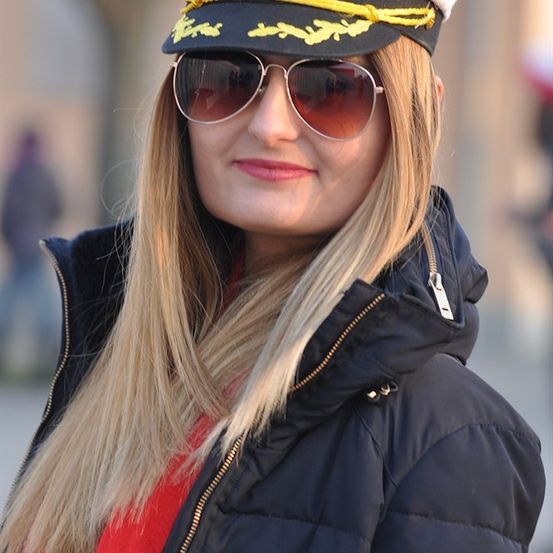Eine Frau mit langen blonden Haaren, die eine dunkelblaue Pufferjacke, ein rotes Shirt und eine dunkelblaue Matrosenmütze mit gelbem Tau trägt. Sie lächelt und trägt eine Sonnenbrille.