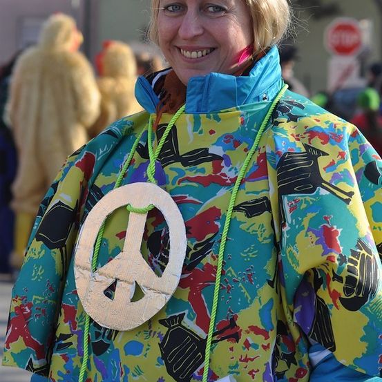 Eine Frau in einem farbenfrohen Friedenszeichen-Jacke lächelt in die Kamera.