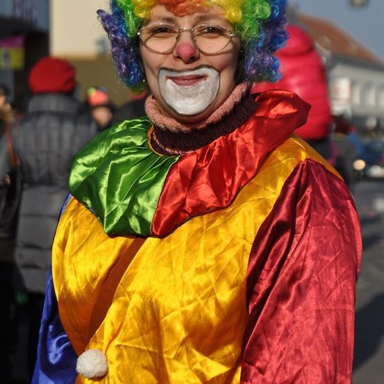 Eine Person in Clown-Verkleidung mit einer bunten Perücke, Brille und bemaltem Lächeln steht auf der Straße.