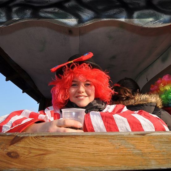 Eine Frau mit roten Haaren in einem Clownkostüm sitzt in einem Fahrzeug und hält einen Plastikbecher.