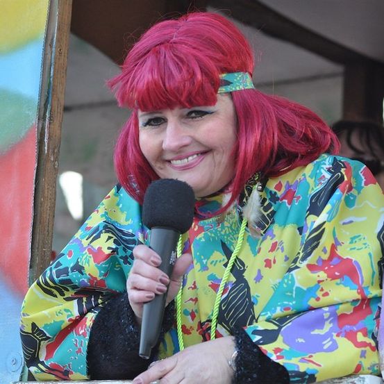 Eine Frau mit roten Haaren, die ein buntes Jackett trägt und ein Mikrofon hält, lächelt und schaut aus einem Fenster.