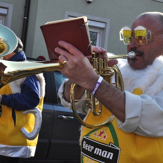 Ein Mann in einem Biermann-Kostüm spielt eine Trompete und hält ein Notenbuch in den Händen. Hinter ihm spielt eine weitere Person in einem ähnlichen Kostüm eine Tuba.
