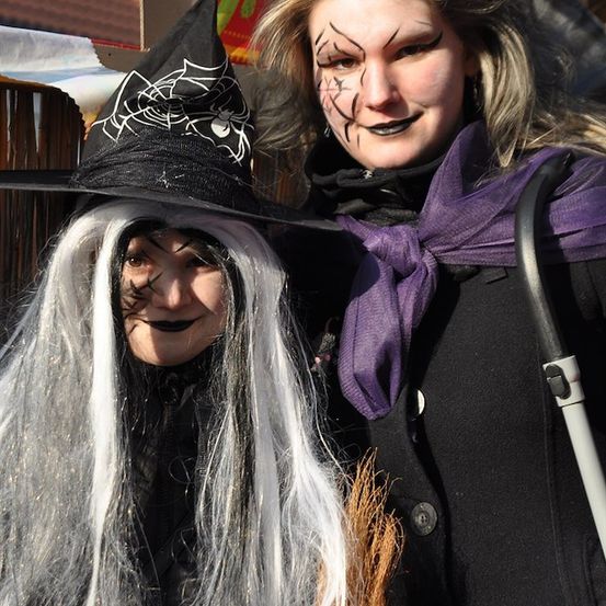 Zwei Frauen in Halloween-Kostümen, eine als Hexe mit schwarzem Hut und Gesichtsbemalung, die andere als Hexe mit einem lila Schal.