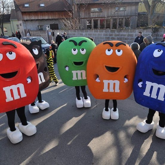 Fünf Personen, verkleidet als M&M-Figuren, stehen in einer Reihe auf einer Straße mit Gebäuden und Bäumen im Hintergrund.