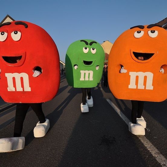 Drei Personen in M&M's-Kostümen, jeweils in Rot, Grün und Orange, gehen die Straße entlang.