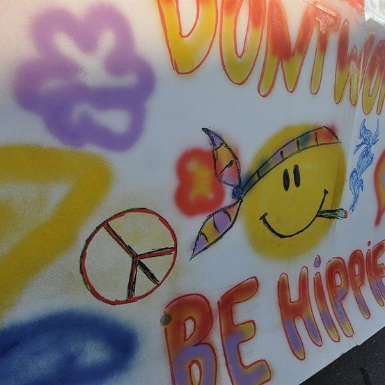Ein farbenfrohes Banner mit einem lächelnden Gesicht, Friedenssymbol und den Worten 'Mach dir keine Sorgen, sei Hippie'. Der Hintergrund ist mit verschiedenen Formen und Farben bemalt.
