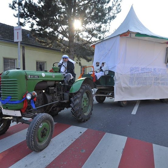 Ein grüner Traktor mit einem weißen Zeltanhänger fährt die Straße entlang. Eine Person sitzt im Fahrersitz, trägt einen weißen Umhang und einen weißen Turban. Eine weitere Person sitzt im Anhänger.