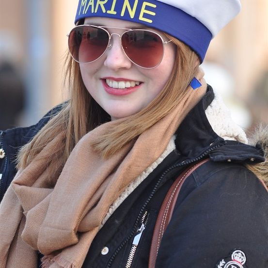 Eine Frau trägt einen weißen und blauen Hut mit dem Wort 'MARINE', eine Sonnenbrille, einen Schal und eine schwarze Jacke mit einem Logo auf der rechten Seite.