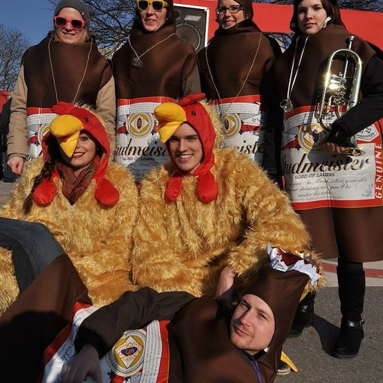 Eine Gruppe von Menschen in Kostümen, die Bierflaschen und Hühnern ähneln, stehen in einer Reihe, wobei eine Person liegt.