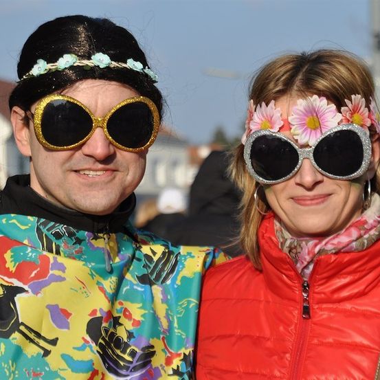 Ein Mann und eine Frau in farbenfrohen Kostümen und mit Blumen auf den Sonnenbrillen lächeln für die Kamera.