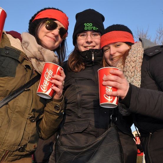 Drei Frauen in Winterkleidung halten rote Coca-Cola-Becher und lächeln für die Kamera.