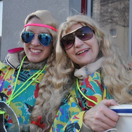 Zwei Frauen in bunten Kostümen, eine hält einen Eimer, beide tragen Sonnenbrillen und lächeln für ein Foto.