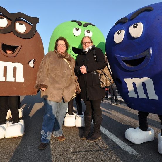 Zwei Frauen stehen zwischen drei M&M's-Mascotten in großen Kostümen, eine braun, eine grün und eine blau, auf einer Straße mit klarem Himmel im Hintergrund.