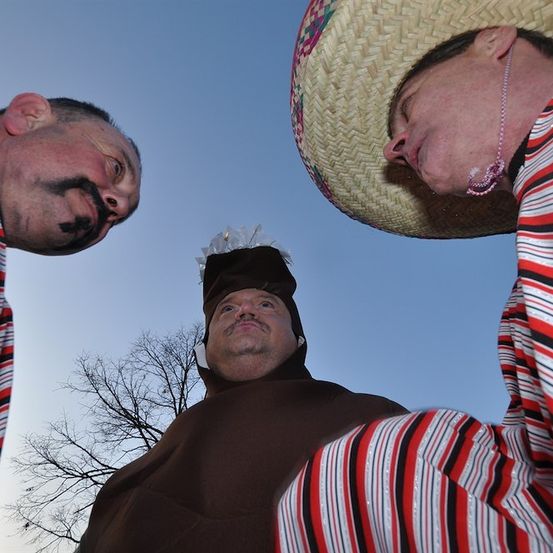 Drei Männer in Kostümen, einer mit einem Sombrero, ein anderer mit einem gemalten Schnurrbart und der dritte in einem braunen Umhang, schauen nach oben.