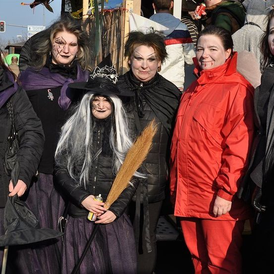 Fünf Frauen in Halloween-Kostümen posieren für ein Foto. Eine Frau hält einen Besen und eine Dose Sprühfarbe. Hinter ihnen gehen Menschen auf einer Straße.